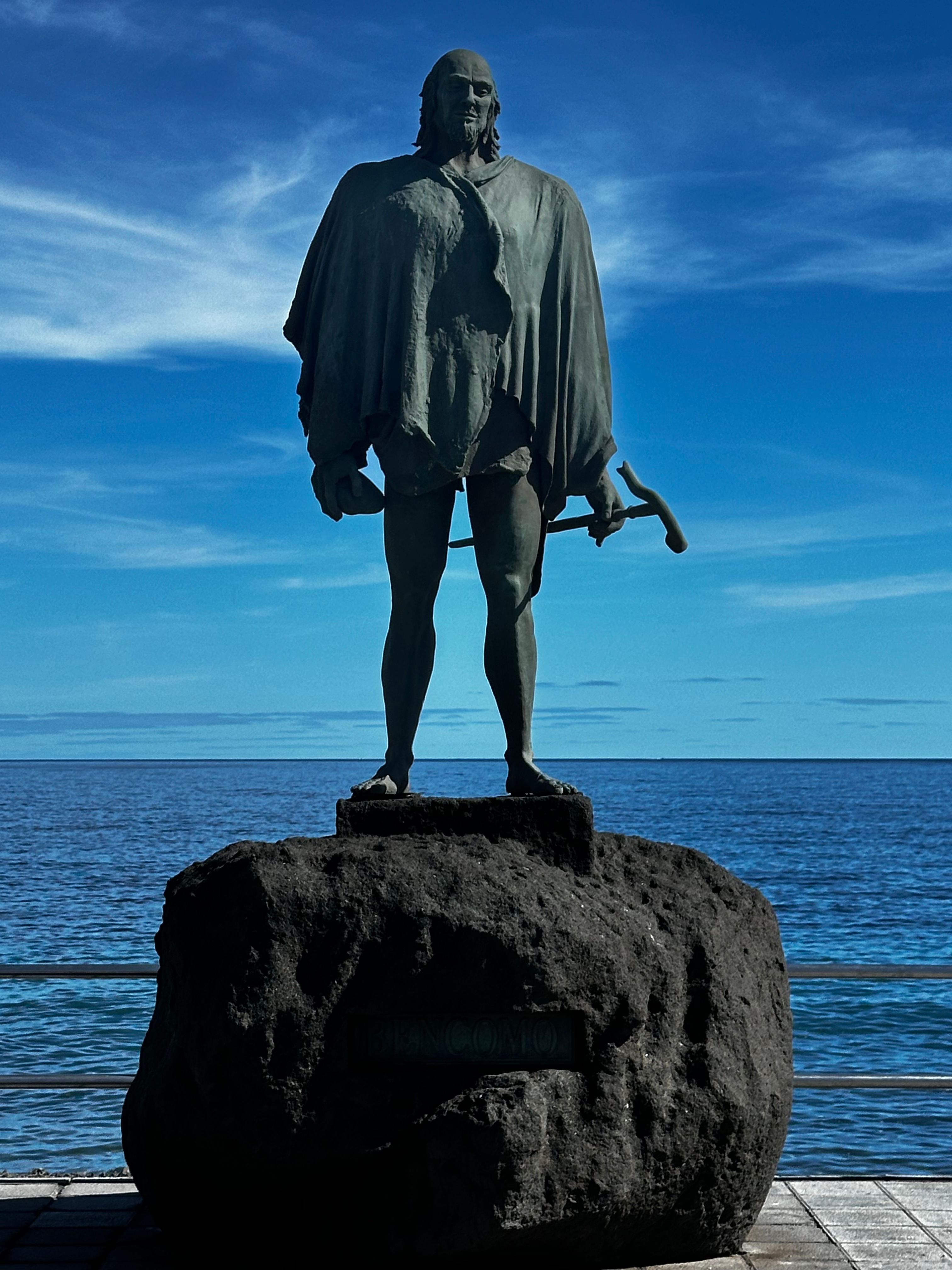 Escultura de bronce del mencey Bencomo sobre un pedestal de roca volcánica, con el mar detrás.