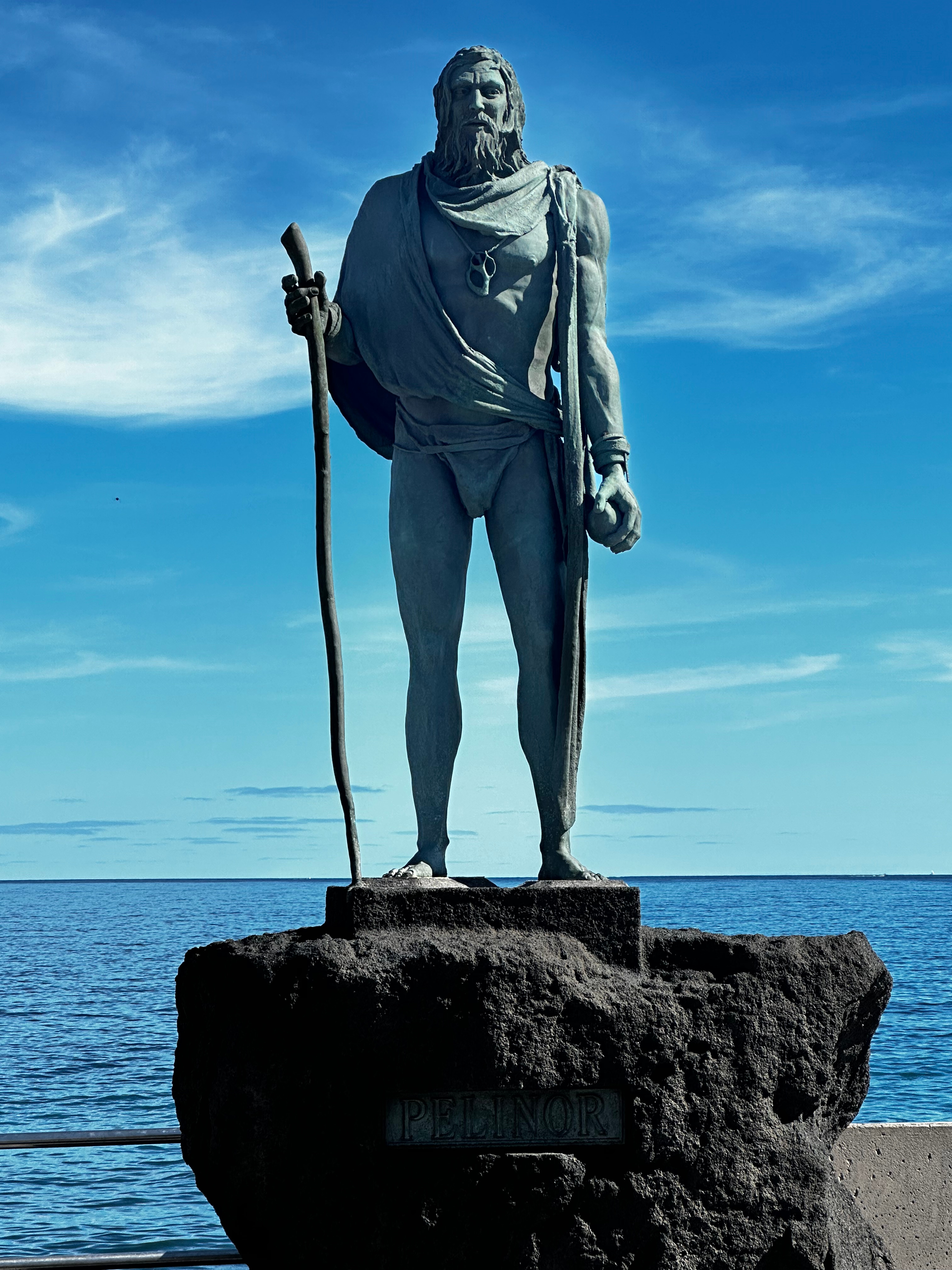 Escultura de bronce del mencey Pelinor con bastón, sobre un pedestal de roca volcánica frente al mar.