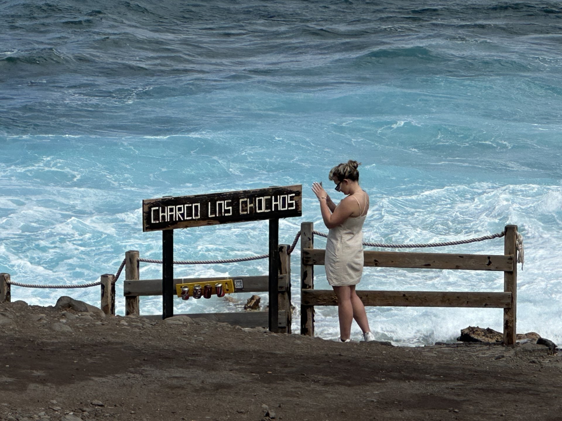 Cartel de madera con el nombre del Charco Los Chochos junto a la barandilla del paseo, con el mar batiendo contra la costa al fondo.