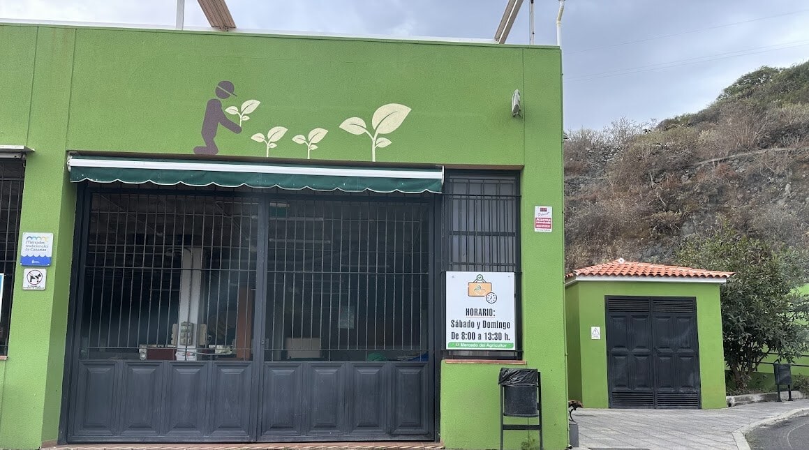Fachada verde del Mercado del Agricultor de La Guancha, con el dibujo de un agricultor plantando brotes y el cartel de horario junto a la reja de entrada.