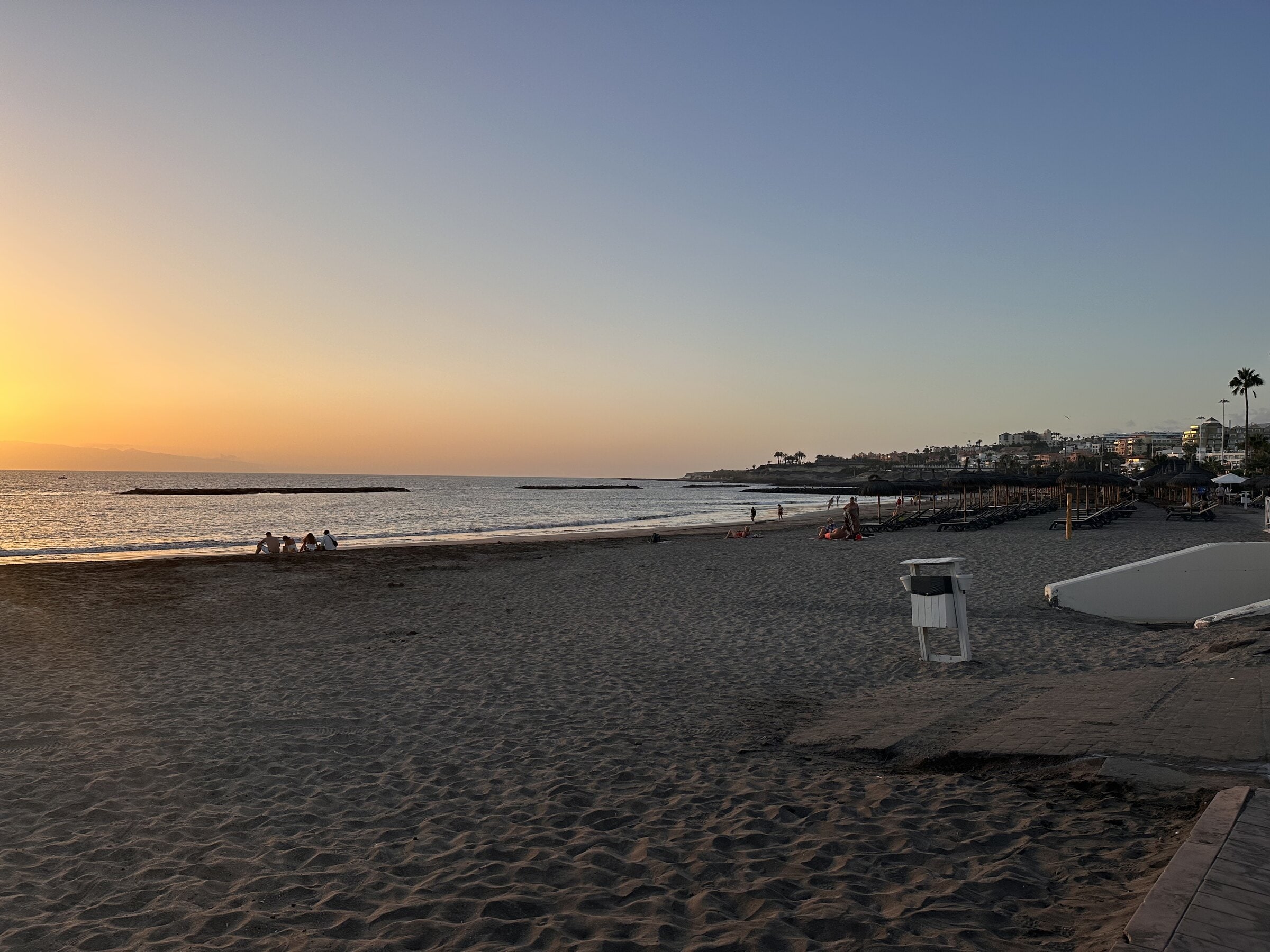 Playa de Torviscas al atardecer, con la arena oscura en primer plano, el sol bajo sobre el mar y la costa de Adeje al fondo.