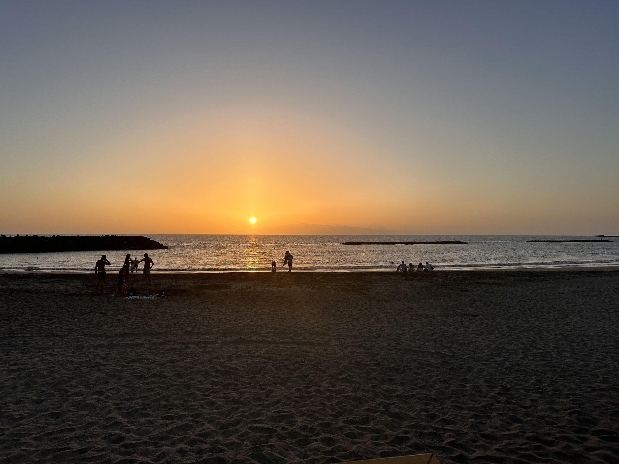 El sol se pone sobre el mar frente a la Playa de Torviscas, con algunas personas sentadas en la orilla a lo lejos.