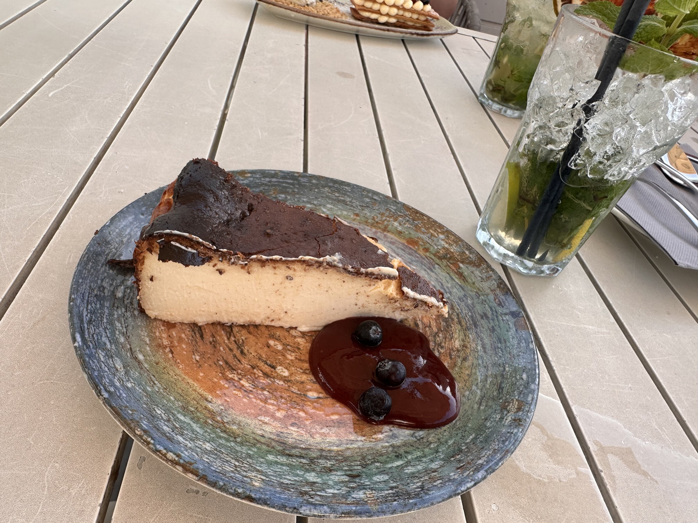 Porción de tarta de queso con salsa de frutos rojos y arándanos, junto a un cóctel con hierbabuena y lima, sobre una mesa de lamas blancas.