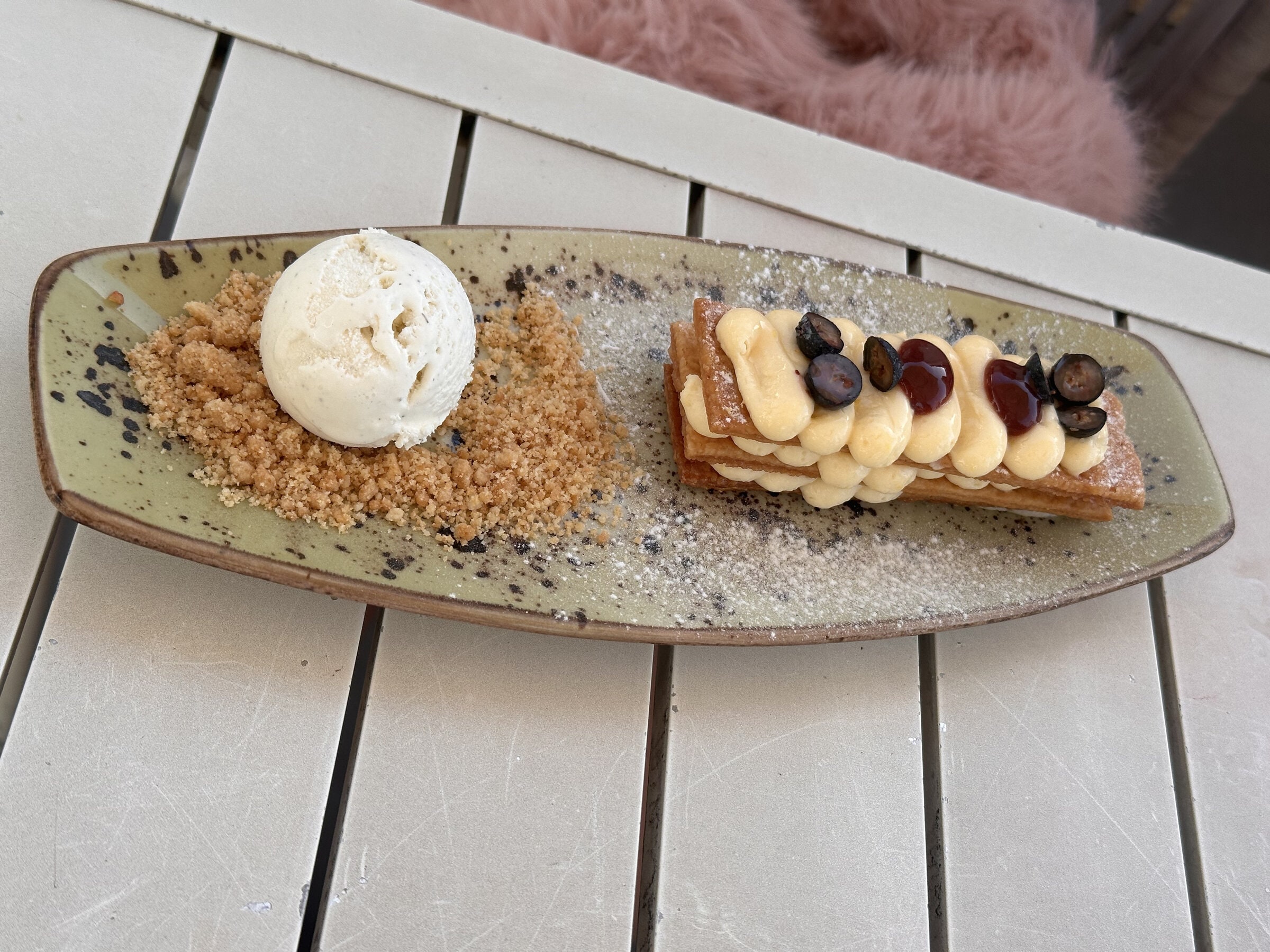 Milhojas de crema con arándanos y una bola de helado sobre crumble, en una fuente alargada.